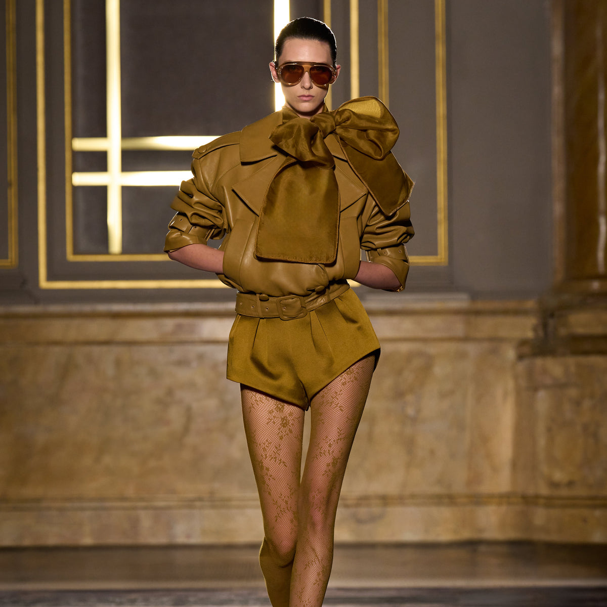 A model walks the runway wearing a structured olive-brown mini dress with dramatic oversized bow details on the shoulders, matching belt, sheer patterned tights, and dark sunglasses in an indoor fashion show setting.