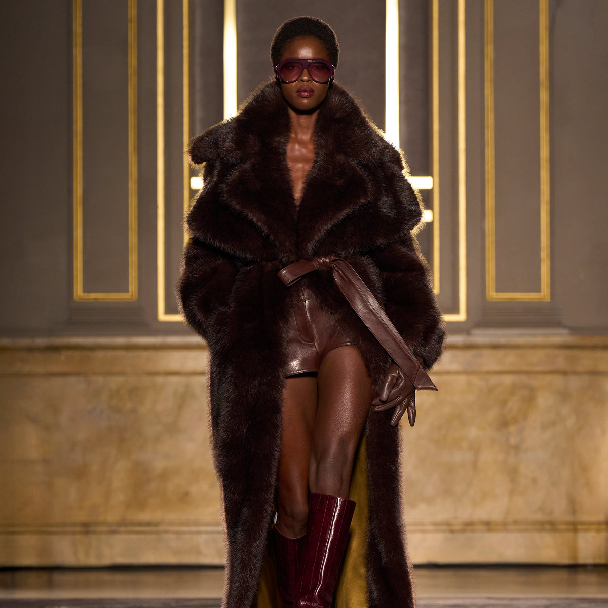 A model walks the runway wearing an oversized dark brown fur coat with a wide collar, a matching belt, sheer brown mini skirt, and glossy burgundy knee-high boots, set against an elegant interior with gold-trimmed gray walls.