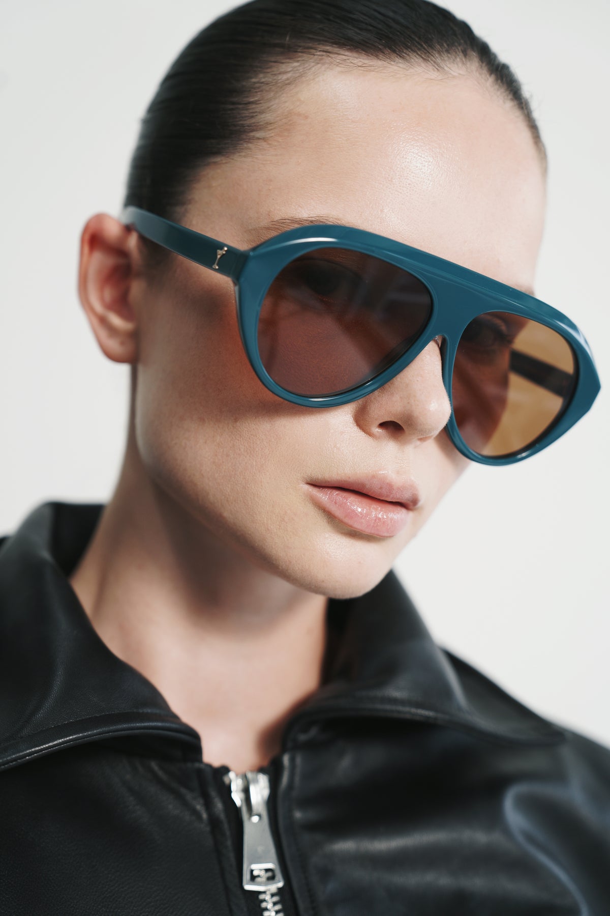 A close-up of a model wearing oversized aviator-style sunglasses with a teal-blue frame and warm brown lenses, paired with a black leather jacket against a light gray background.