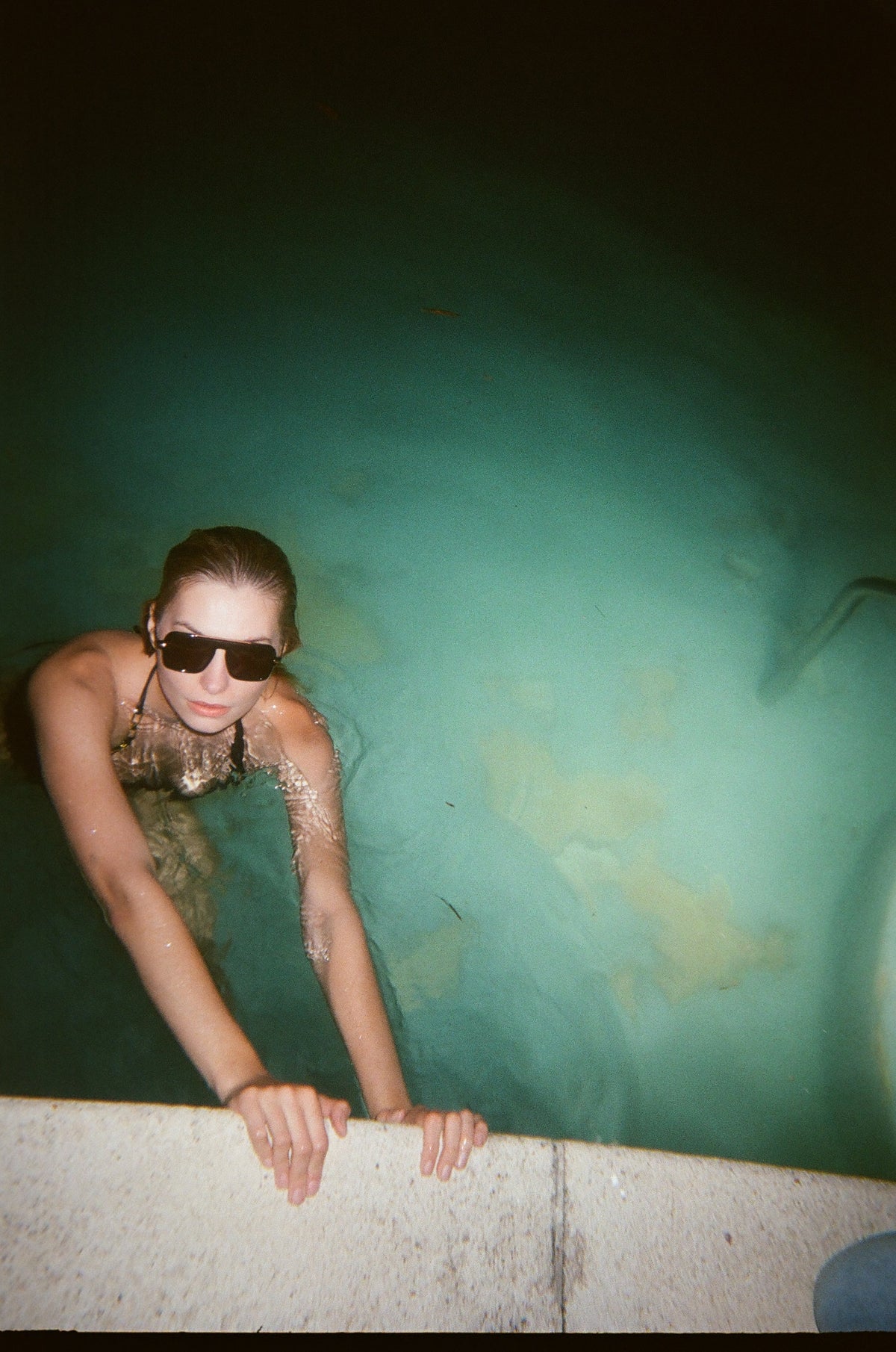 Woman in a green-lit pool at night wearing Past Midnight black sunglasses, film photograph