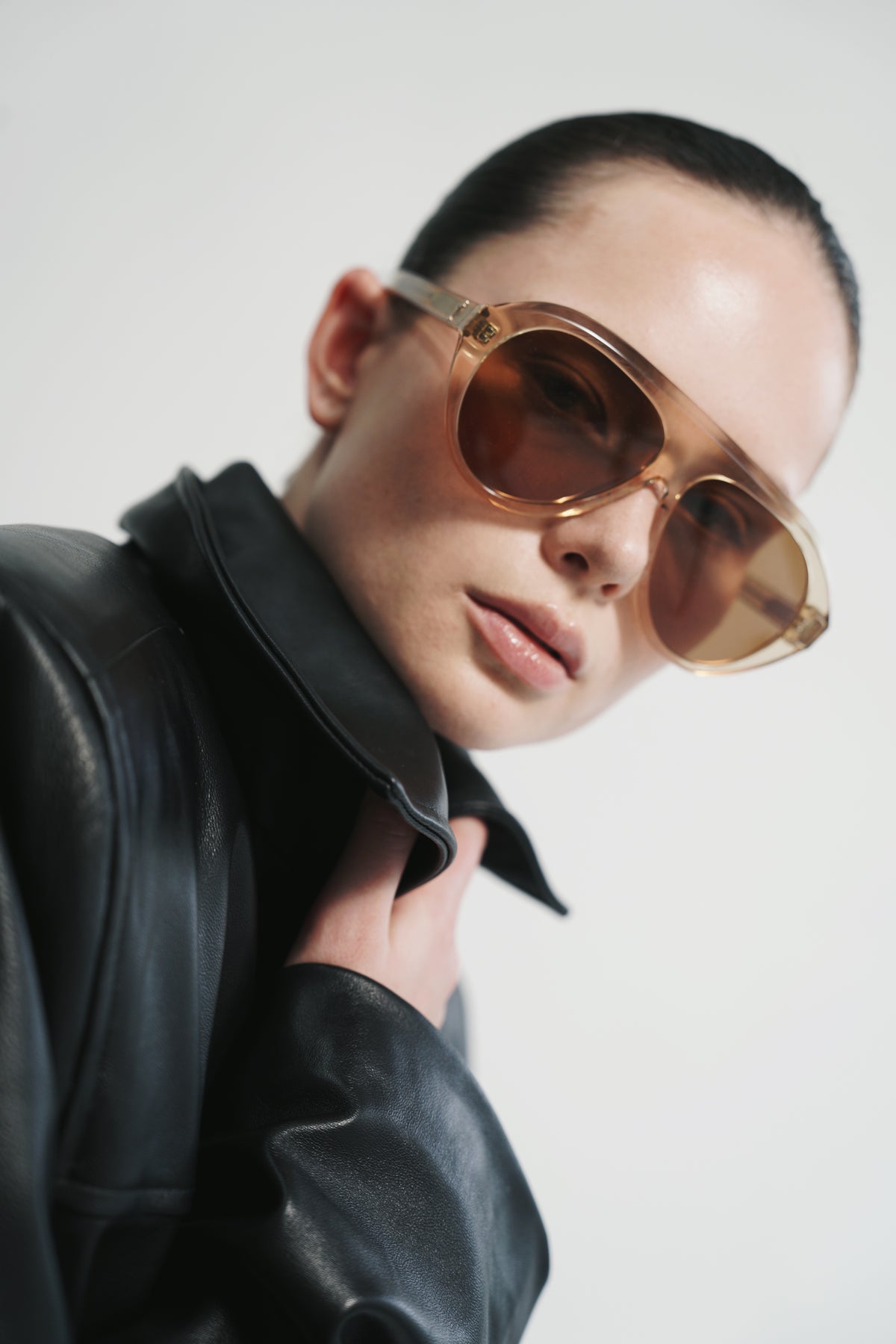 A close-up portrait of a model wearing oversized aviator-style sunglasses with translucent amber-tinted lenses and gold-toned frames, paired with a black leather jacket against a plain light background.
