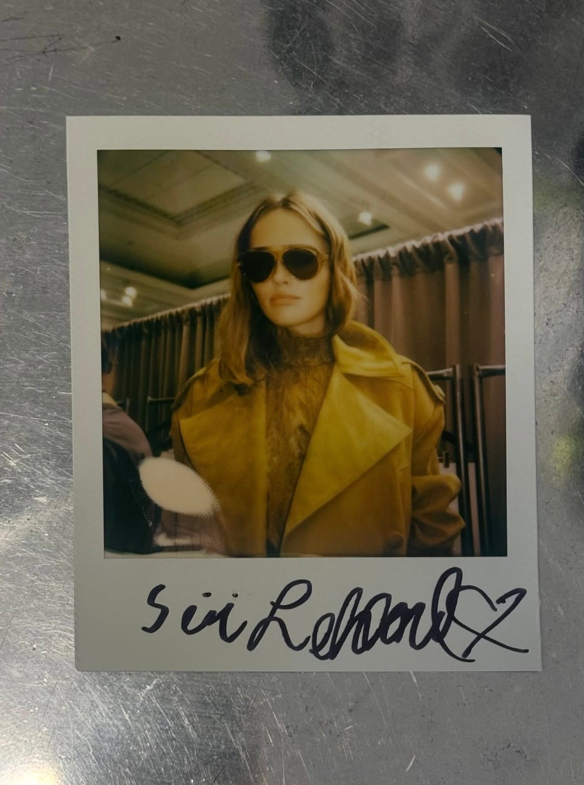 An autographed Polaroid-style photo print featuring a woman in a mustard yellow coat and dark sunglasses, placed on a scratched metallic surface. The signature at the bottom reads 'sui Romanz'.