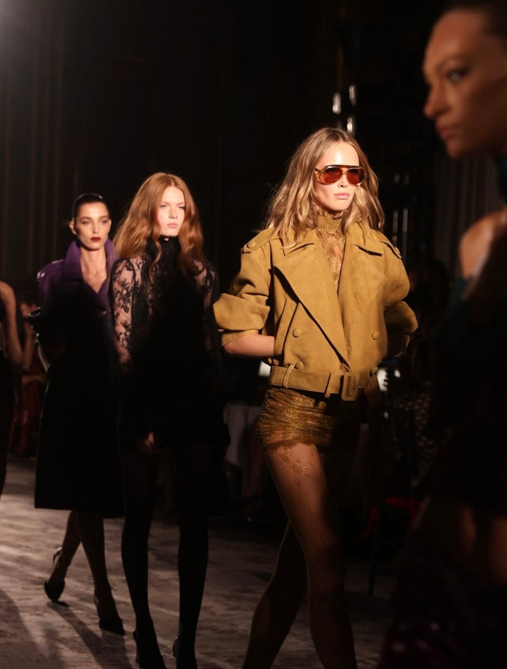 A runway fashion photo showing models walking on a catwalk. The central model wears a tan cropped jacket, a metallic gold mini skirt, and orange-tinted sunglasses, while other models in dark clothing walk behind her in dim lighting.