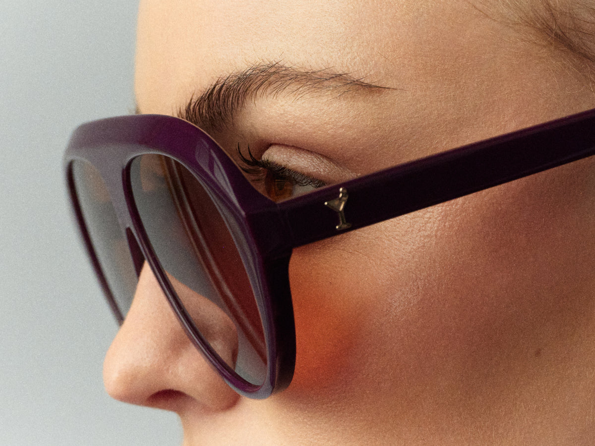 Close-up of a person wearing oversized dark purple sunglasses with dark lenses and a small gold logo on the temple arm, against a light gray background.