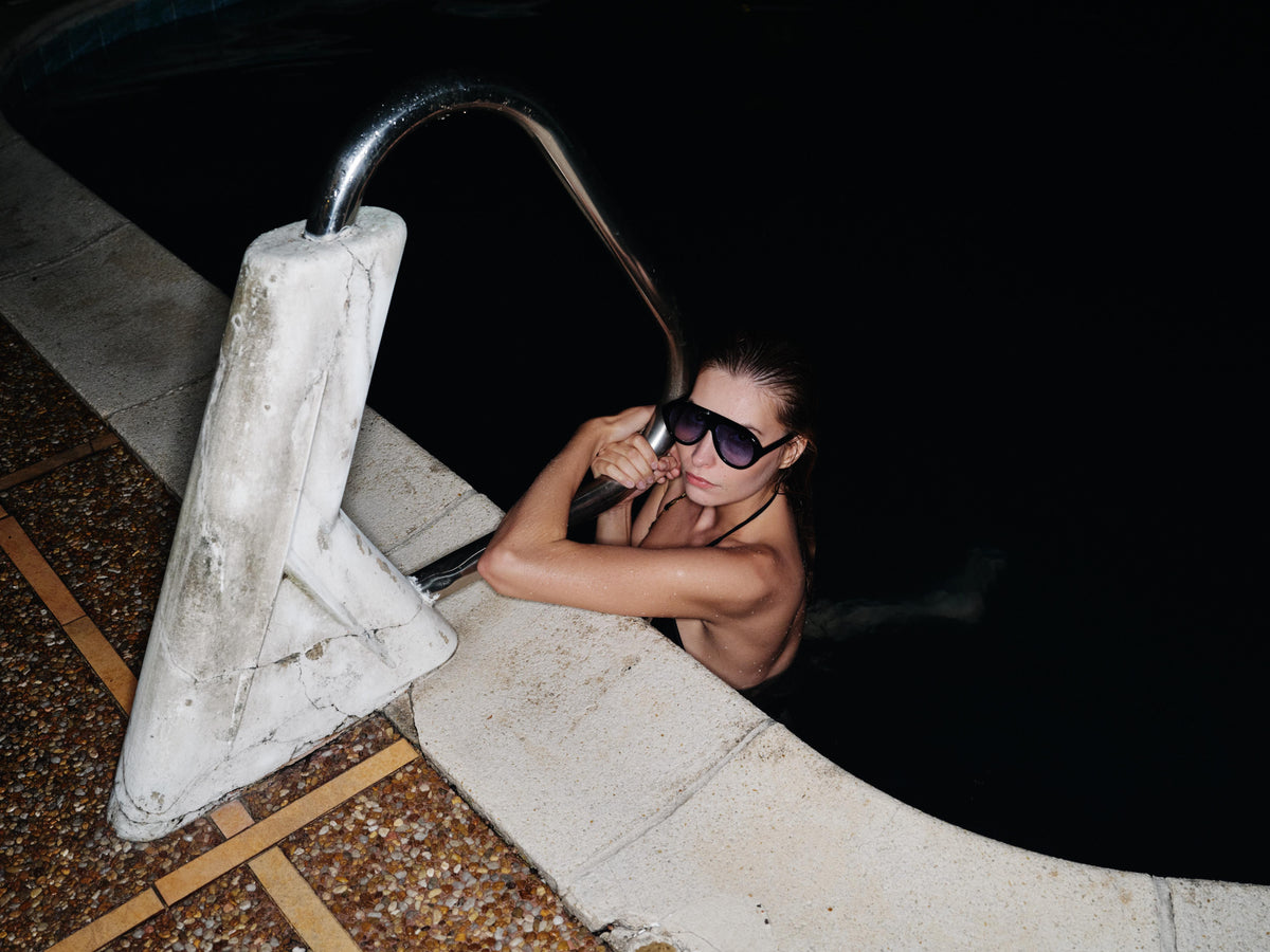 Woman by a sculptural pool ladder at night wearing Past Midnight The Icon Dark Blue shield sunglasses with dark blue lenses
